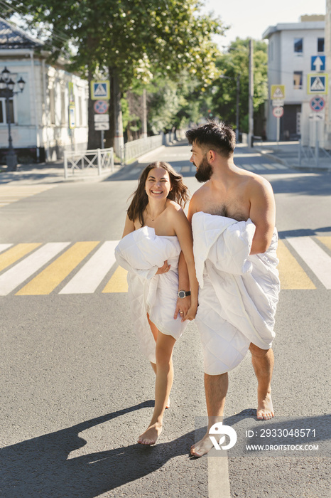 Young couple in love, a man and a woman, are running along the road in blankets at dawn.