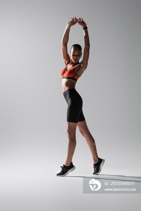 full length of tattooed african american woman in sports bra and shorts stretching with raised hands on grey.