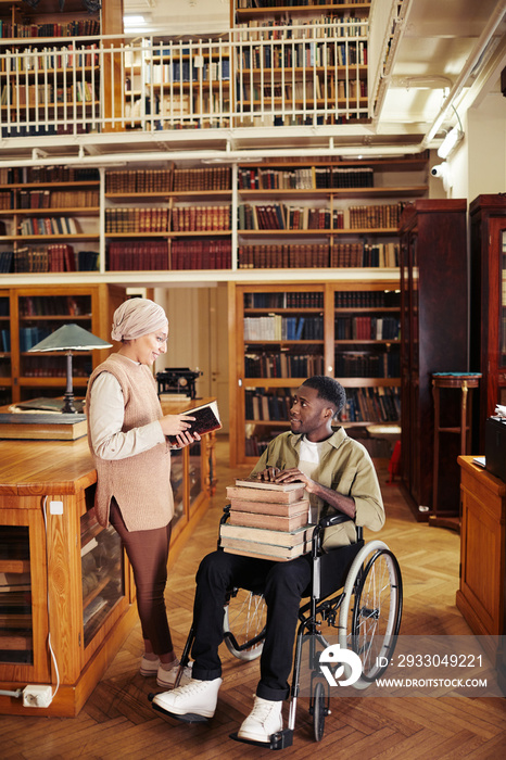 Full length portrait of young African-American man in wheelchair holding stack of books and chatting with friend at college library