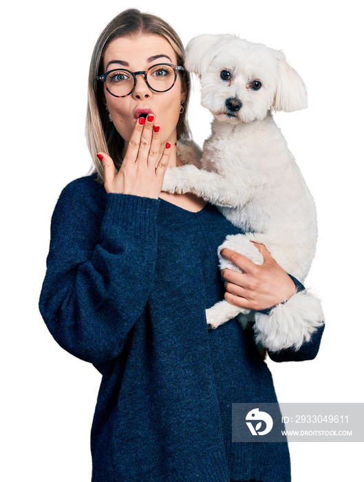 Young blonde woman hugging cute dog covering mouth with hand, shocked and afraid for mistake. surprised expression