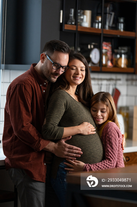 Affectionate young Caucasian family embracing at home while standing in the kitchen. Touching mom’s pregnant belly.