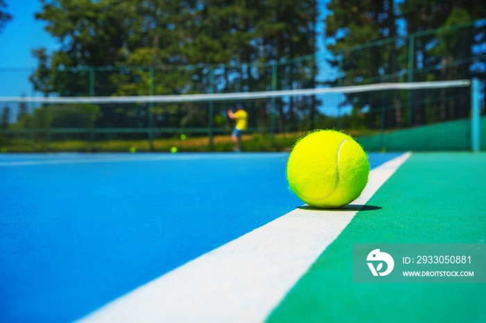 Tennis ball on white court line on hard modern blue green court with player, net, balls, trees on the background. Close-up, selective focus. Sport, tennis play, healthy lifestyle concept.