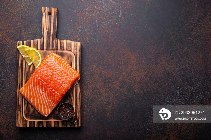 Fresh raw salmon fillet on wooden kitchen cutting board with seasonings and lemon top view on dark brown rustic background, space for text