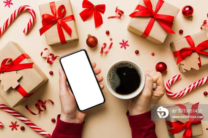 Above view of female hand holding smart phone with hot cup of coffee. Christmas decoration on colored background