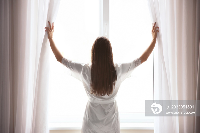 Young woman opening curtains at home