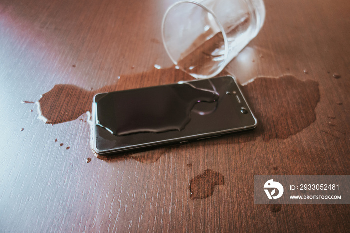 Smartphone wet by accident on wooden table. Glass of water spilled.