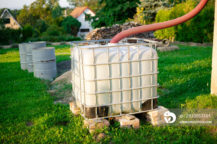 Device for collecting rainwater used for watering plants on organic farms. Big white plastic water tank is elevated from the ground on brick pillars. Rain barrels in the allotment garden