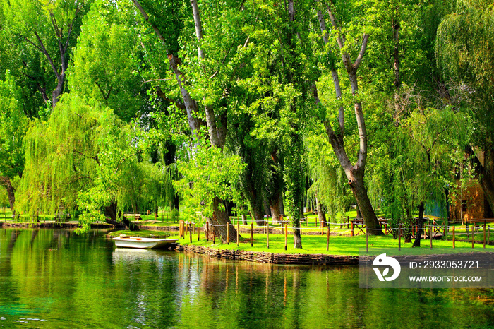 Wonderful view at the quiet water surface of a pond reflecting fresh greenery of various trees and a small boat moored to the shore of a neat green island in Fonti del Clitunno park