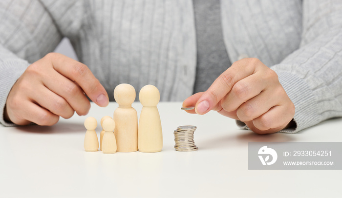 female hand puts coins in a pile and wooden figurines of a family on a white table. Economy and investment concept, budget planning. Receiving subsidies from the state, tax