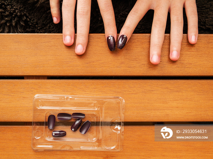 Female attaching fake nails on a wooden table, Cosmetic glamor procedure. Top down view. Spare nails in a plastic container