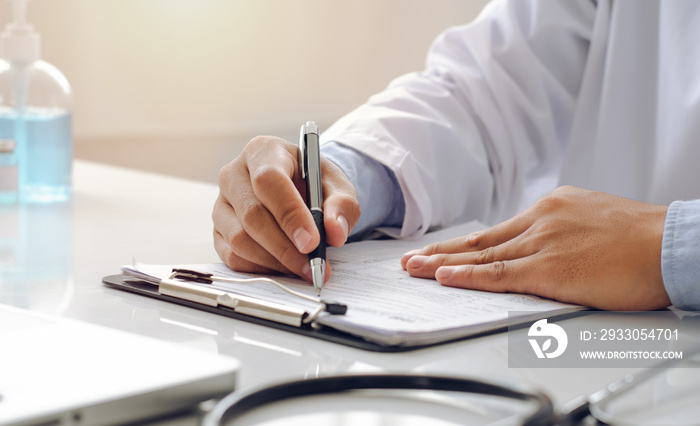 A male doctor with a uniform sits at a table in the doctor’s office and writes a prescription on a piece of paper.