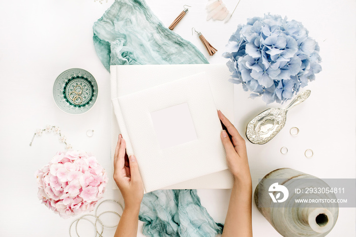 Woman hands hold family wedding photo album, pastel colorful hydrangea flower bouquet, turquoise blanket, decoration, fashion accessories on white background. Flat lay, top view.