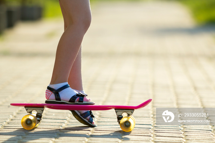 Child slim legs in white socks and black sandals on plastic pink skateboard on bright sunny summer blurred copy space pavement background. Outdoors activities and healthy lifestyle concept.