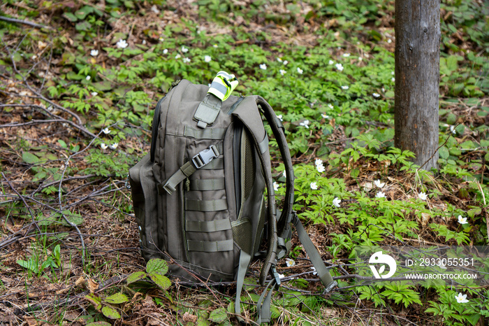 Tactical military bag on the ground in the forest. Backpack for hiking and hunting. Army bag on green grass background near tree and flowers. Camouflage backpack suitable for the forest.