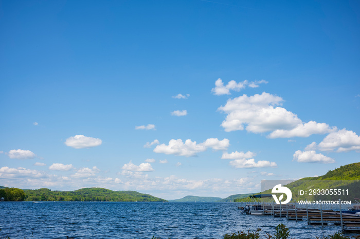 The blue waters of Otsego Lake in Cooperstown, New York,  on a sunny summer day with cumulus clouds in the sky, photographed near a dock.