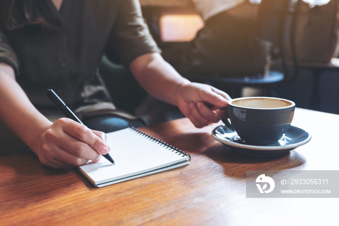 Closeup image of a woman’s hand writing down on a white blank notebook while drinking coffee on wooden table