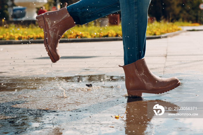 Woman wearing rain rubber boots walking running and jumping into puddle with water splash and drops in autumn rain.