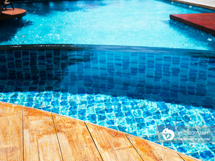 Empty brown wooden plank table or deck floor curved shape in front of the blurred background of blue mosaic tiles grid pattern in large swimming pool. Empty space on poolside, summer background.