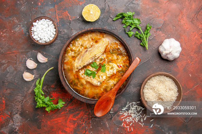 top view homemade chicken and rice soup in a bowl wooden spoon bowls with sea salt and rice garlic parsley leaves slice of lemon on dark red background