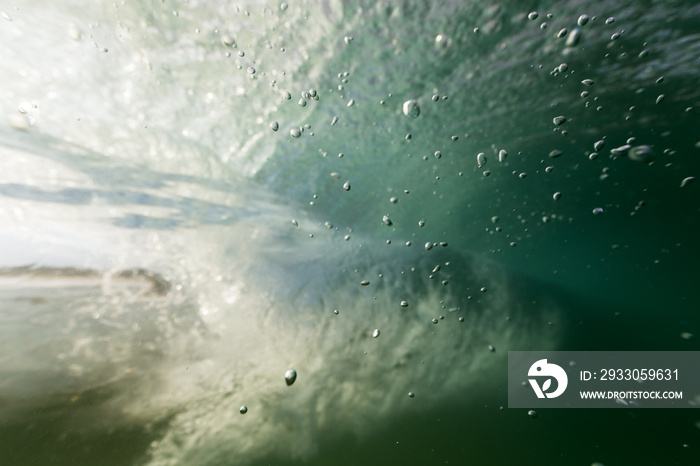 Bubbles rise in clear ocean water from the sea floor in this underwater shot as a breaking wave passes in the background.