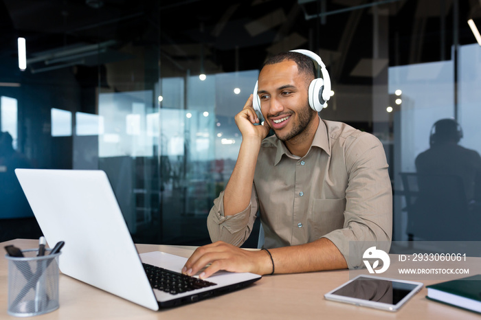 Young hispanic businessman in shirt working inside office using laptop at work, man with headphones listening to music and audio podcasts at work.