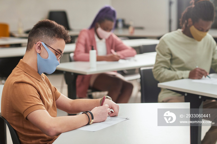 Side view portrait of young man wearing mask while taking test or exam in school with diverse group of people, copy space