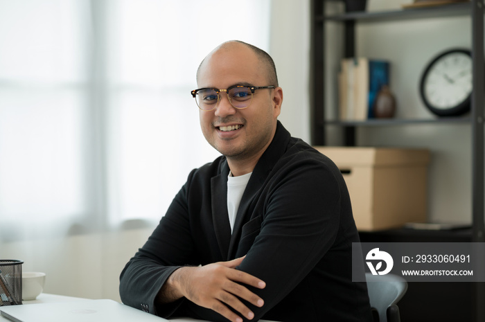 A middle-aged man around the age of 35. Working at home looking at camera. He was wearing a black suit and glasses. Smiling asian businessman work from home.