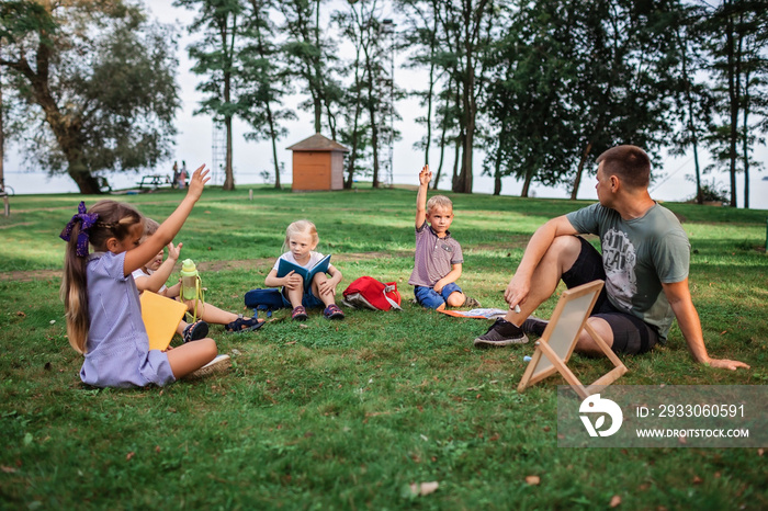 Back to school. Kindergarten and elementary scholars sitting with teacher on grass at open-air class