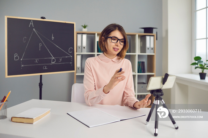 Remote teaching. College professor or school math teacher having online class, using mobile phone with tripod. Smart young woman in glasses explaining geometry problem in virtual lesson via video call