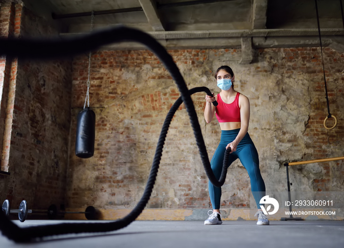 Young woman exercising with battle ropes at the gym. Strong female athlete doing crossfit workout with battle rope during epidemic COVID-19