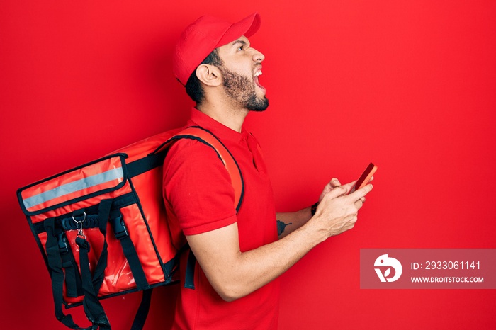 Hispanic man with beard wearing delivery package backpack using smartphone angry and mad screaming frustrated and furious, shouting with anger looking up.