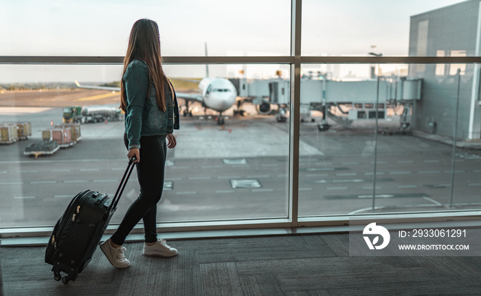 Young traveler woman looking at plane in airport glass window, waiting for flight, tourist near luggage in departure lounge. Travel Concept