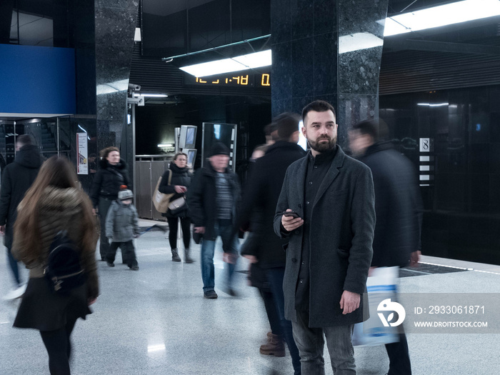 Handsome bearded man dressed in wool coat holds a smartphone and stands still in metro station within moving crowds of people