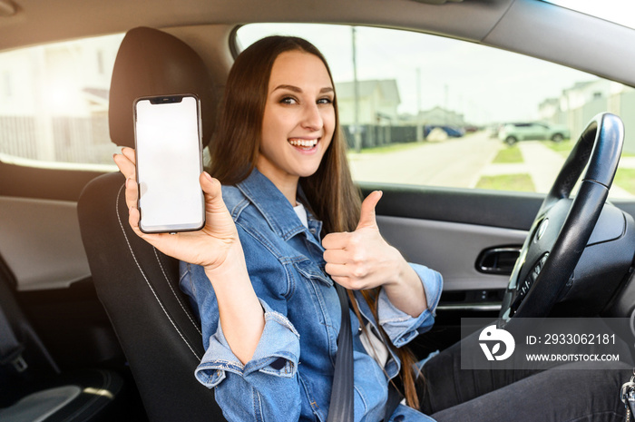 A young happy woman driving a car, she shows empty phone screen and thumb up. Useful mobile apps for driver. Side view inside car