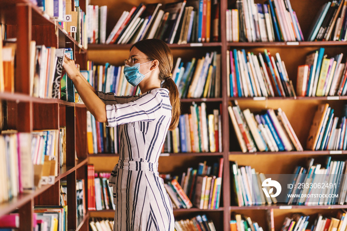 Young attractive librarian with face mask on searching for a book while standing in library during corona virus pandemic.