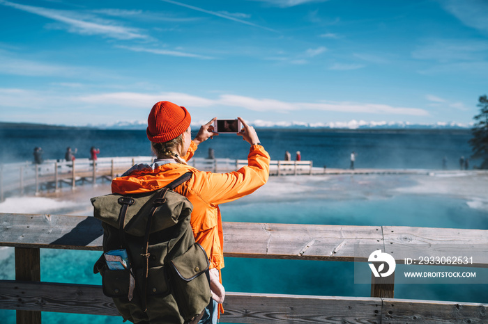 Back view of female traveler using smartphone camera for taking picture of hot springs, hipster girl with backpack making photo getting to touristic landmark in Yellowstone during american trip