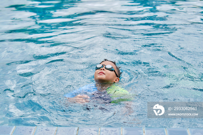 Cute little Asian 6 years old school boy child in swimming goggles learning to swim, floating, treading water to stay afloat at outdoor pool in summer day, Happy kid having fun on summer vacation