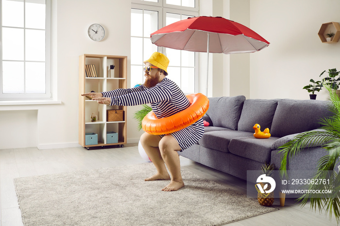 Funny chubby guy staying at home and having cool fake beach holiday in living room. Happy plus size man in hat and striped swimsuit pretends to jump in imaginary ocean waters. Summer vacation concept