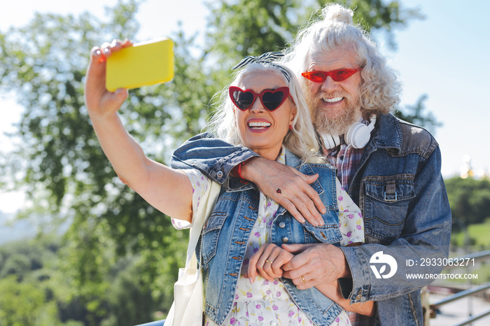 Modern device. Nice cheerful woman holding her smartphone while taking a selfie together with her husband