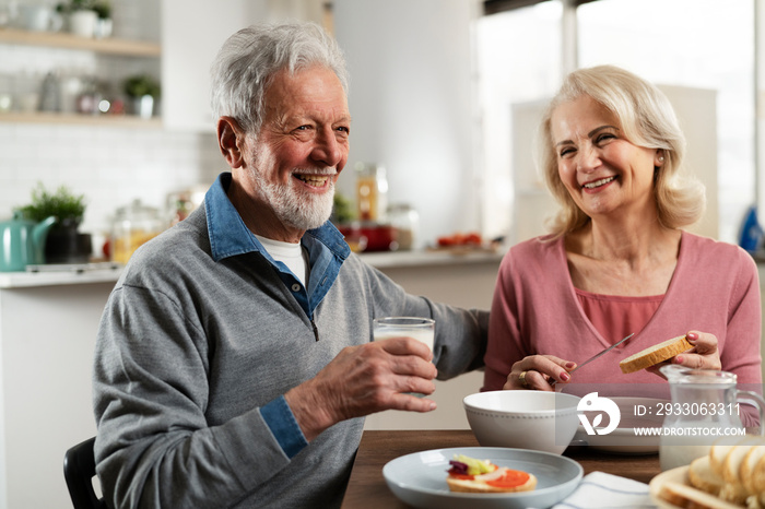 Senior couple eating breakfast in the kitchen. Husband and wife talking and laughing while eating a sandwich.