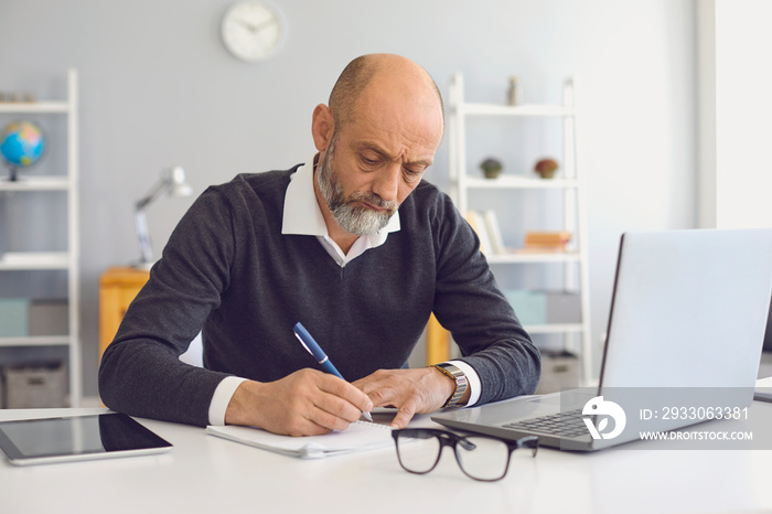 Working and studying from home. Thoughtful mature man taking notes during online business meeting or webinar