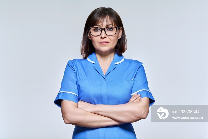 Portrait of middle aged nurse in blue uniform looking at camera on light studio background