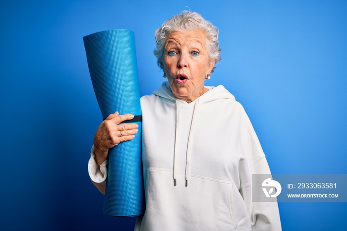 Senior beautiful sporty woman holding mat for yoga standing over isolated blue background scared in shock with a surprise face, afraid and excited with fear expression