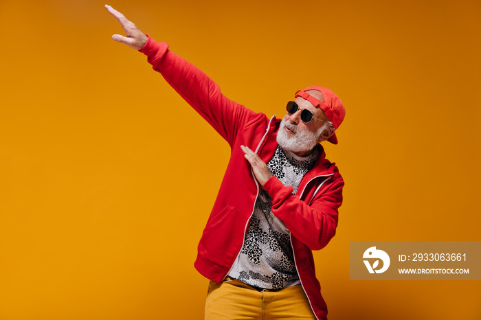 White bearded man in sunglasses and cap posing on orange background. Cool guy in red hoodie and leopard print t-shirt has fun..