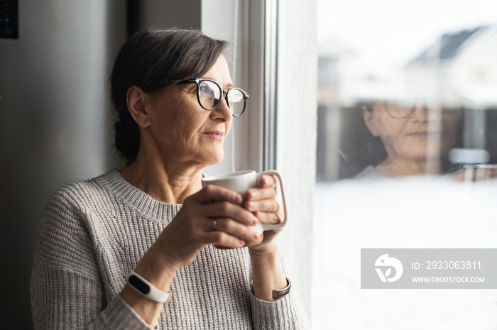 Close-up portrait of senior older woman wearing glasses enjoys morning coffee in the kitchen at home. A modern retirement lady daydreaming with a mug of hot drink looks through the window