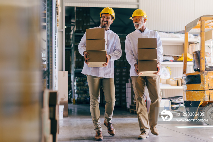 Two smiling workers in white uniforms and with helmets on heads carrying boxes in storage.