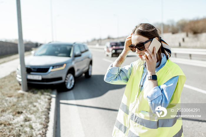 Embarrassed woman calling road assistance standing near the car during the road accident on the highway