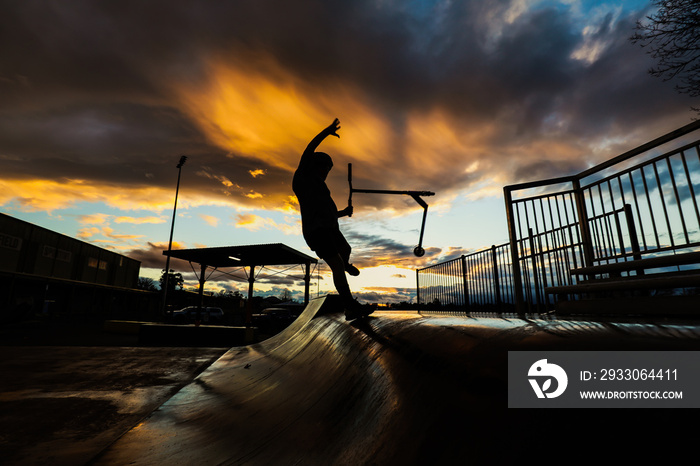 Silhouette image of boy riding scooter at skate park jumping ramp at sunset