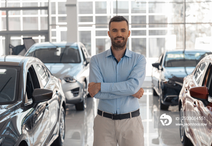 Young male consultant in auto show standing near cars and looking at camera