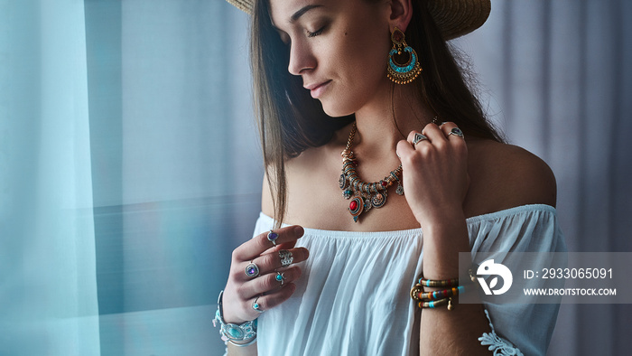 Stylish brunette boho chic woman wears white blouse and straw hat with big earrings, bracelets, golden necklace and silver rings. Fashionable hippie gypsy bohemian outfit with jewelry details